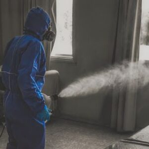 Person in protective suit and mask spraying pesticide indoors for pest control treatment.