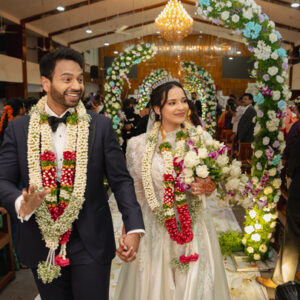 A smiling bride and groom, both wearing floral garlands, walk hand-in-hand down the aisle under a floral arch after their wedding ceremony.